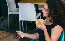 Вид сбоку на счастливую молодую латиноамериканку, просматривающую мобильный телефон и наслаждающуюся вкусным капучино за столом уличного кафе — стоковое фото