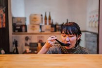 Young Asian woman in sweater eating ramen with spoon at wooden counter in cafe — Stock Photo