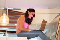 Sorridente freelance femminile navigando netbook mentre seduto nelle scale a casa con luce al neon rosa e lavorando al progetto — Foto stock