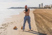 Hombre calvo y sin camisa de cuerpo completo mirando a la cámara haciendo columpios con kettlebell mientras está parado descalzo en la costa de arena con la ciudad en el fondo - foto de stock
