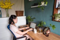 Vista lateral de la mujer asiática freelancer leer documentos en el ordenador portátil mientras está sentado en la mesa durante el trabajo remoto - foto de stock