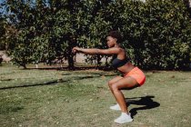 Side view of young fitness African American woman doing squats in the park on a sunny day — Stock Photo