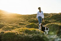 Cuerpo completo de mujer étnica feliz con perro Border Collie caminando juntos por el sendero entre colinas cubiertas de hierba en la soleada noche de primavera - foto de stock