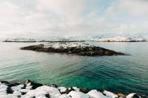 Кам'яні острівці, розташовані в хвилястому морі біля снігового гірського хребта проти хмарного неба взимку на Лофотенських островах, Норвегія. — стокове фото