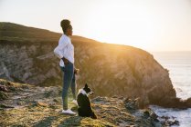 Vista lateral da jovem dona afro-americana com o cão Border Collie passando tempo juntos na praia perto do mar acenando ao pôr do sol contemplando vistas — Fotografia de Stock