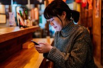 Side view of satisfied Asian lady in casual sweater smiling while using mobile phone at counter in traditional ramen bar — Stock Photo