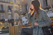 Вид сбоку современной миллениалки в стильном весеннем наряде, сидящей на скамейке и отвечающей на телефонный звонок, пока она отдыхает на городской улице, глядя в сторону — стоковое фото