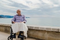 Старший чоловік в інвалідному візку на набережній дивиться в роздумах і насолоджується видом на море — стокове фото