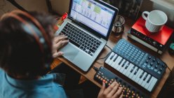 Зверху невідомий зосереджений молодий чоловік в навушниках, що працюють на синтезаторі та ноутбуці за столом вдома — стокове фото