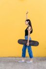 Seitenansicht einer jungen Skaterin mit erhobenem Arm und Skateboard, die tagsüber auf dem Gehweg mit bunter gelber Wand im Hintergrund wegschaut — Stockfoto