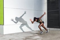 Seitenansicht einer afroamerikanischen Athletin, die vorwärts springt, während sie in der Nähe einer modernen Hauswand auf der Stadtstraße läuft — Stockfoto