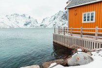 Casa de campo amarela e cais nevado localizado perto do mar ondulante contra montanhas no dia frio de inverno na aldeia costeira nas Ilhas Lofoten, Noruega — Fotografia de Stock