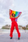 Von unten eine stylische Afroamerikanerin in trendiger Kleidung, die Flagge mit Regenbogenornament hisst, während sie in die Kamera auf der Fahrbahn blickt — Stockfoto
