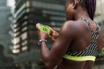 Unerkennbare Afroamerikanerin surft Fitness-App auf Smartphone, während sie beim Outdoor-Training auf verschwommenem Hintergrund der Stadtstraße steht — Stockfoto