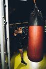 Young focused Asian man training boxing performing punches while exercising with heavy punching bag in a modern gym — Stock Photo