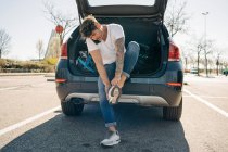 Tätowierte männliche Sportler ziehen Schuhe an, während sie mit dem Handy gegen Auto mit offenem Kofferraum in der Stadt reden — Stockfoto