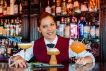 Усміхнена жінка-банкір стоїть за барною стійкою з різними видами алкогольних напоїв, що подаються в творчих коктейльних келихах у формі гриба та риби — стокове фото