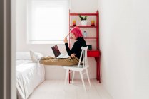Vista lateral de una joven freelancer enfocada en ropa casual sentada en una silla y usando un portátil mientras trabaja en un proyecto en un apartamento moderno y luminoso - foto de stock