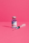 Флакон коронавирусной вакцины возле шприца с иглой на розовом фоне — стоковое фото