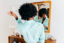Vista posterior de una hermosa joven afroamericana en traje casual tocando el pelo rizado mientras está de pie en la habitación y mirando el espejo - foto de stock