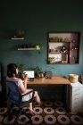 Вид сбоку азиатской женщины-фрилансера, читающей документы на ноутбуке, сидя и попивая чай за столом во время удаленной работы — стоковое фото