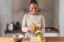 Contenuto Donna afroamericana in abbigliamento con decoro a strisce peeling banane da cucina a tavola in casa — Foto stock