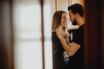 Hombre barbudo alegre abrazando a la mujer sonriente mientras se apoya en el armario con fregadero en la acogedora cocina en casa - foto de stock