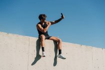 Adatta atleta maschile in abiti sportivi scattare selfie con il cellulare mentre si riposa sulla recinzione dopo l'allenamento — Foto stock