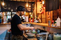 Vista lateral do homem em bandana de pé no balcão e cozinhar ramen no moderno café asiático — Fotografia de Stock