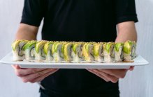 Crop anonymous male waiter standing with plate of delicious sushi rolls topped with soy sauce and sesame seeds — Stock Photo