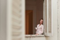 Femme tranquille en pyjama debout près de la fenêtre avec une tasse de café du matin et regardant loin — Photo de stock
