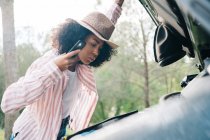 Seitenansicht einer jungen afroamerikanischen Reisenden, die ihr Smartphone mit glühender Taschenlampe hält, während sie den Automotor überprüft, weil er während der Fahrt durch die Natur Probleme hat — Stockfoto