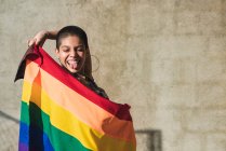 Contenuto giovane donna etnica bisessuale con bandiera multicolore che rappresenta i simboli LGBTQ che guardano dall'alto in basso nella giornata di sole — Foto stock