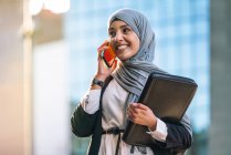 Content Muslim female entrepreneur in hijab and with folder standing in street and speaking on mobile phone while discussing business project — Stock Photo