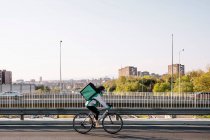 Side view of female courier with thermal bag riding bike on bridge while delivering food in city, copy space above — Stock Photo