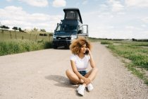 Preocupada joven afroamericana sentada con el pelo rizado hablando por teléfono móvil mientras pide ayuda al servicio de reparación después de un accidente con una caravana en el campo - foto de stock