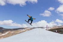 Anonymous male athlete in fabric mask jumping with snowboard over snow against Sierra Nevada and cableway in Spain — Stock Photo
