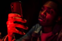 Vista lateral de la cosecha afroamericana calma chico serio en chaqueta de mezclilla llamando en el teléfono móvil en neón misteriosa luz roja sobre fondo negro - foto de stock