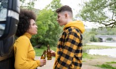 Side view of multiracial young man and woman clinking bottles of beer and looking at each other while relaxing in nature on summer day — Stock Photo