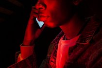 Вид сбоку урожая афро-американский спокойный серьезный парень в джинсовой куртке звонит по мобильному телефону в неоновом загадочном красном свете на черном фоне — стоковое фото