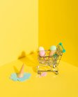 From above composition of miniature shopping trolley with assorted multicolored ice cream cones placed near fallen melting blue ice cream on yellow background — Stock Photo