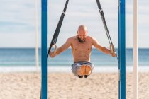 Hombre barbudo sin camisa colgando de anillos gimnásticos con las piernas levantadas entrenamiento duro en la playa de arena mirando hacia abajo - foto de stock