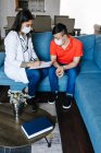 Fröhliche Ärztin im Arztkittel spricht mit Teenie-Jungen mit Down-Syndrom und macht sich Notizen auf Klemmbrett beim Termin zu Hause — Stockfoto