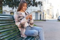 Modern millennial female in stylish spring outfit sitting on bench and browsing on mobile phone while resting on urban street looking away — Stock Photo