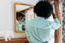 Vue arrière d'une belle jeune femme afro-américaine en tenue décontractée touchant les cheveux bouclés tout en se tenant dans la chambre et en regardant le miroir — Photo de stock