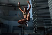 Vista lateral da mulher negra forte pulando alto perto da parede de vidro do edifício moderno enquanto se exercita na rua da cidade no dia ensolarado — Fotografia de Stock