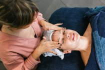 Зверху врожаю невизначений косметолог наносить засоби для чищення обличчя на обличчя жіночого клієнта під час догляду за шкірою в салоні краси — стокове фото