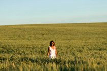 Улыбающаяся молодая черная леди в белом летнем платье прогуливается по зеленому пшеничному полю, глядя в камеру днем под голубым небом — стоковое фото