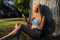 Mulher despreocupada no smartphone e ouvindo música nos fones de ouvido enquanto sentada debaixo da árvore no parque ao pôr do sol no verão — Fotografia de Stock