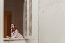 Donna tranquilla in pigiama in piedi vicino alla finestra e parlando sul telefono cellulare a casa — Foto stock